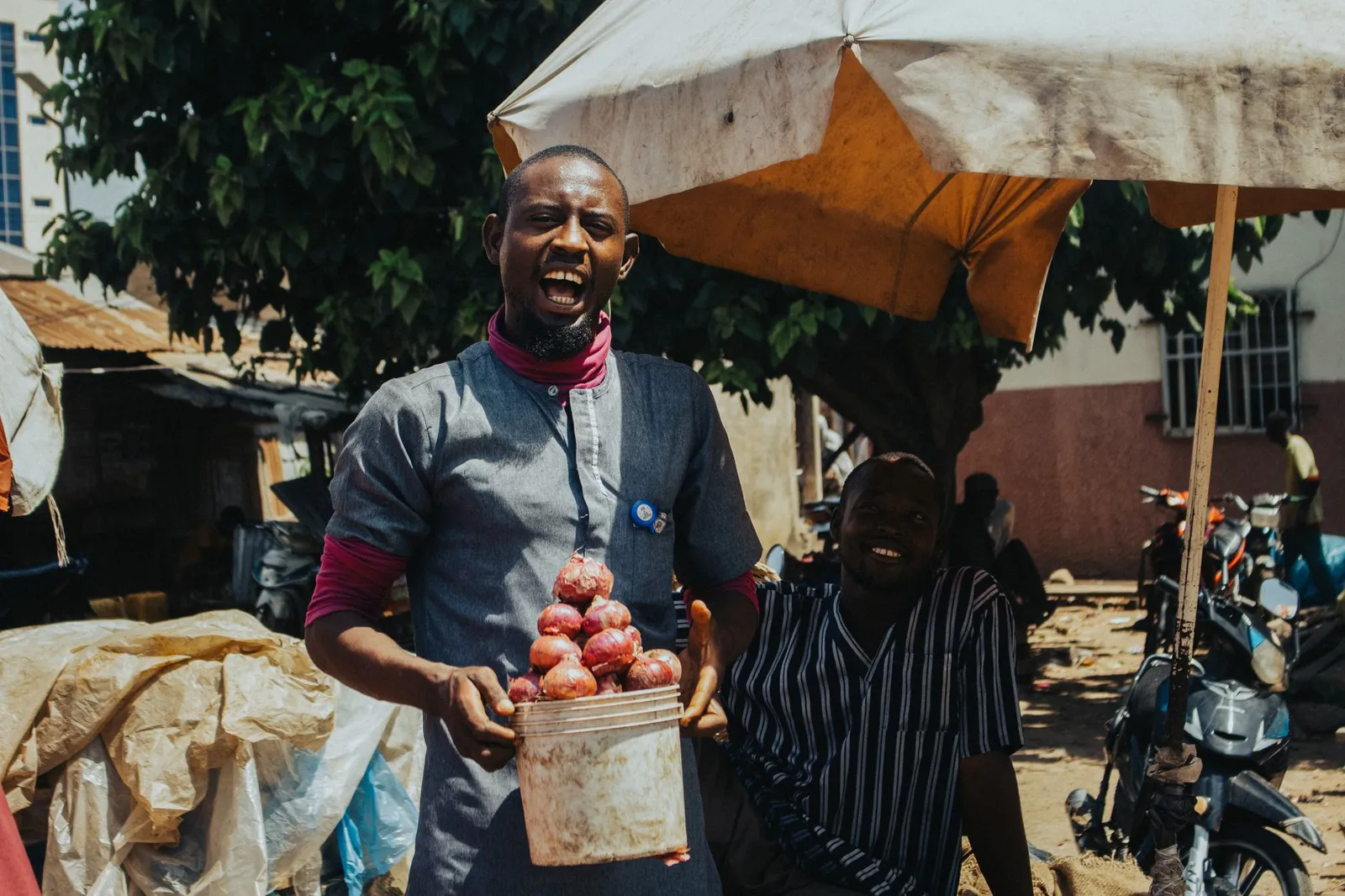 A smiling street trader holding out a bowl of fresh tomatoes under a market sunshade.