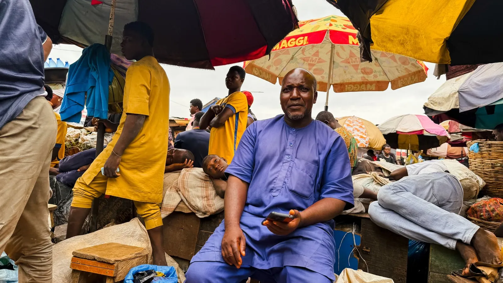 A trader sitting among his goods at a busy Nigerian market, under bright umbrellas.